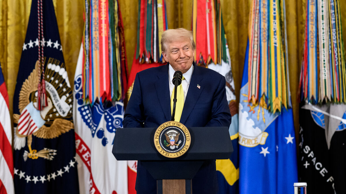 Donald Trump speaking at a podium with US presidential seal, flags in background, referencing Alaska and Russia confusion before Putin meeting