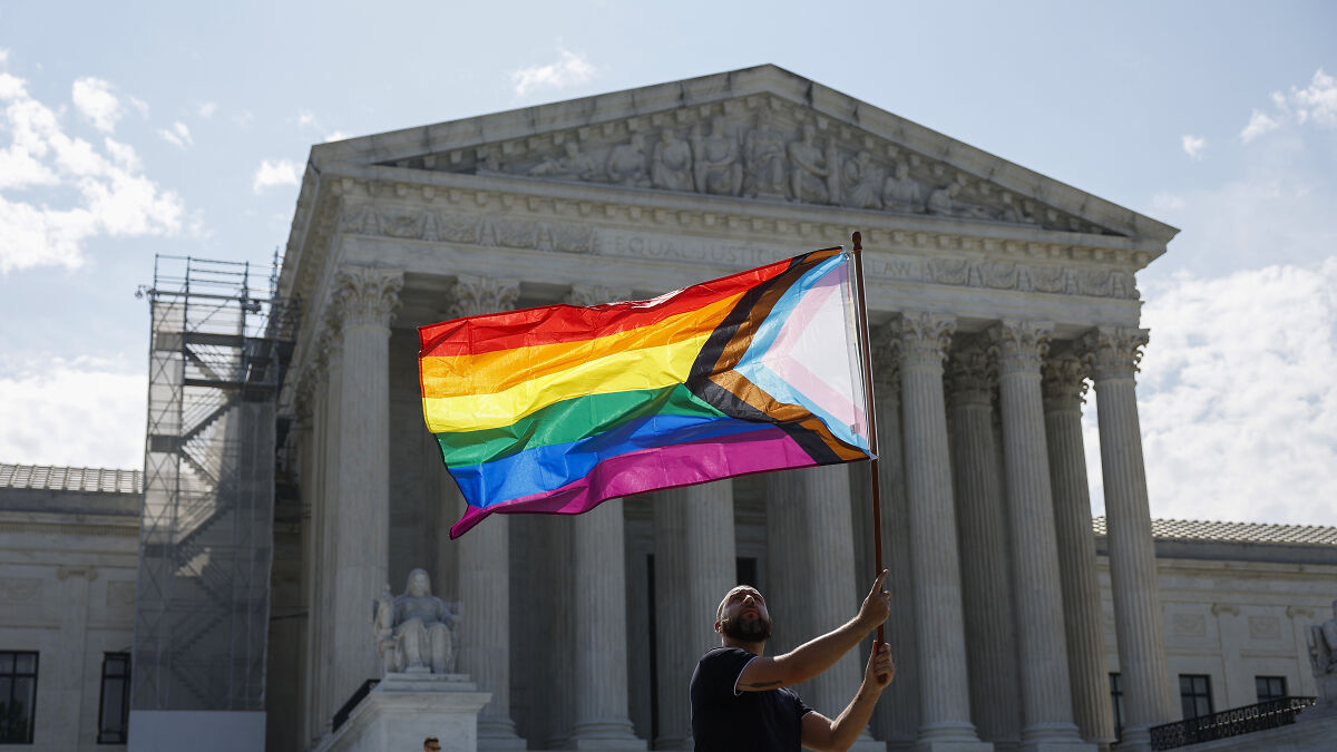Person waving LGBTQ pride flag outside Supreme Court building amid same-sex marriage battle debate.