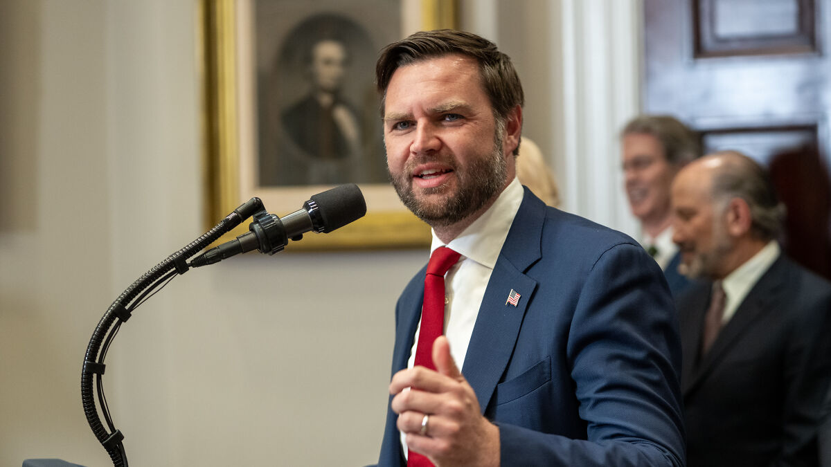 JD Vance speaking at a podium wearing a blue suit and red tie during a political event indoors.