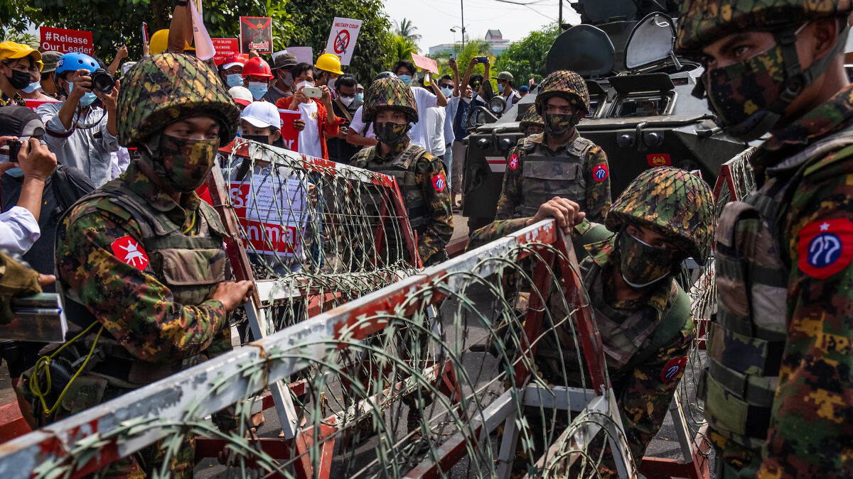 Myanmar junta soldiers in camouflage and masks securing barricades with barbed wire amid protesting civilians.