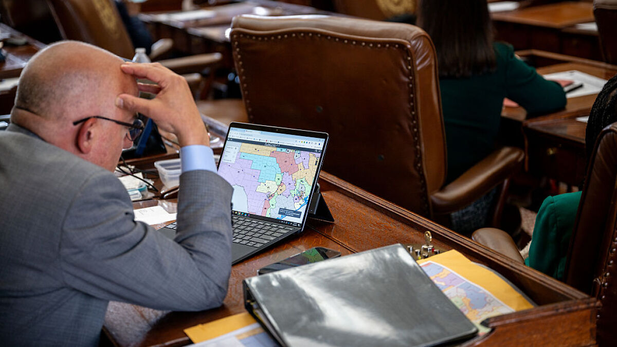 Man in suit reviewing a partisan map on a laptop during Texas GOP redistricting session in a legislative chamber.