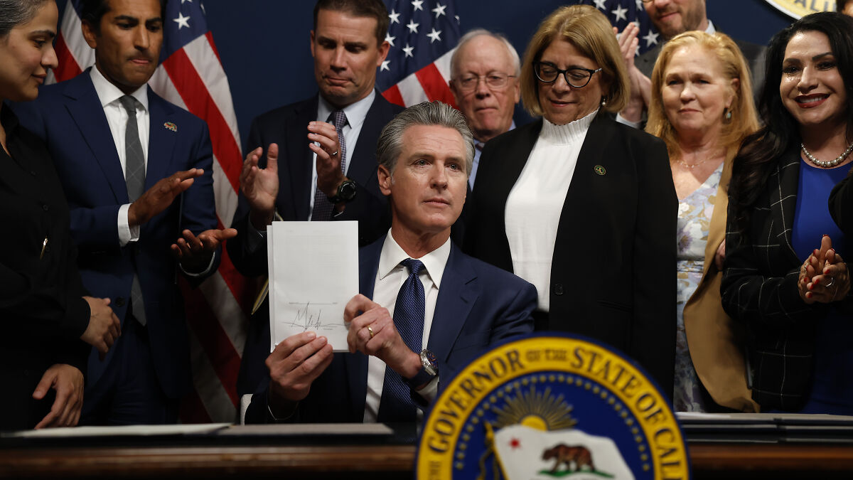 California Governor Newsom holding signed redistricting plan surrounded by applauding officials and American flags in background