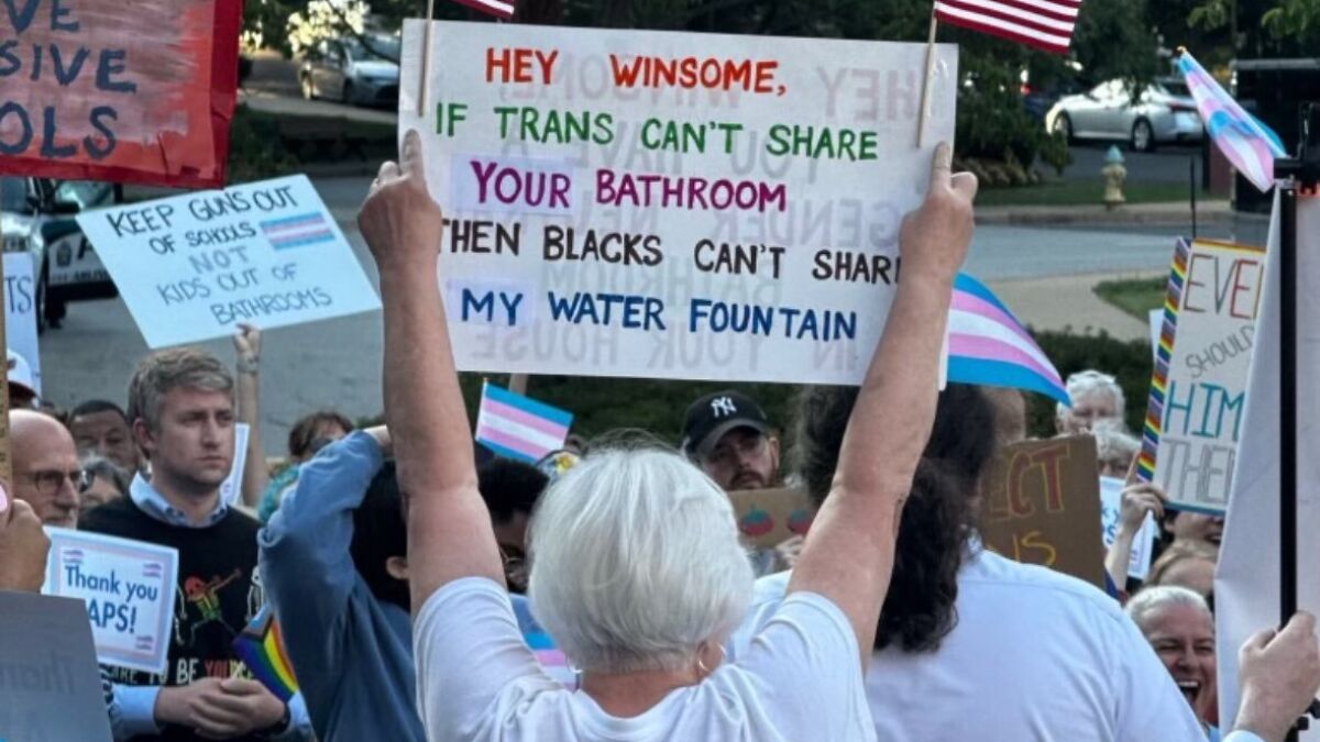 Protester holding racist sign about water fountain at trans rights rally with crowd and transgender flags in background