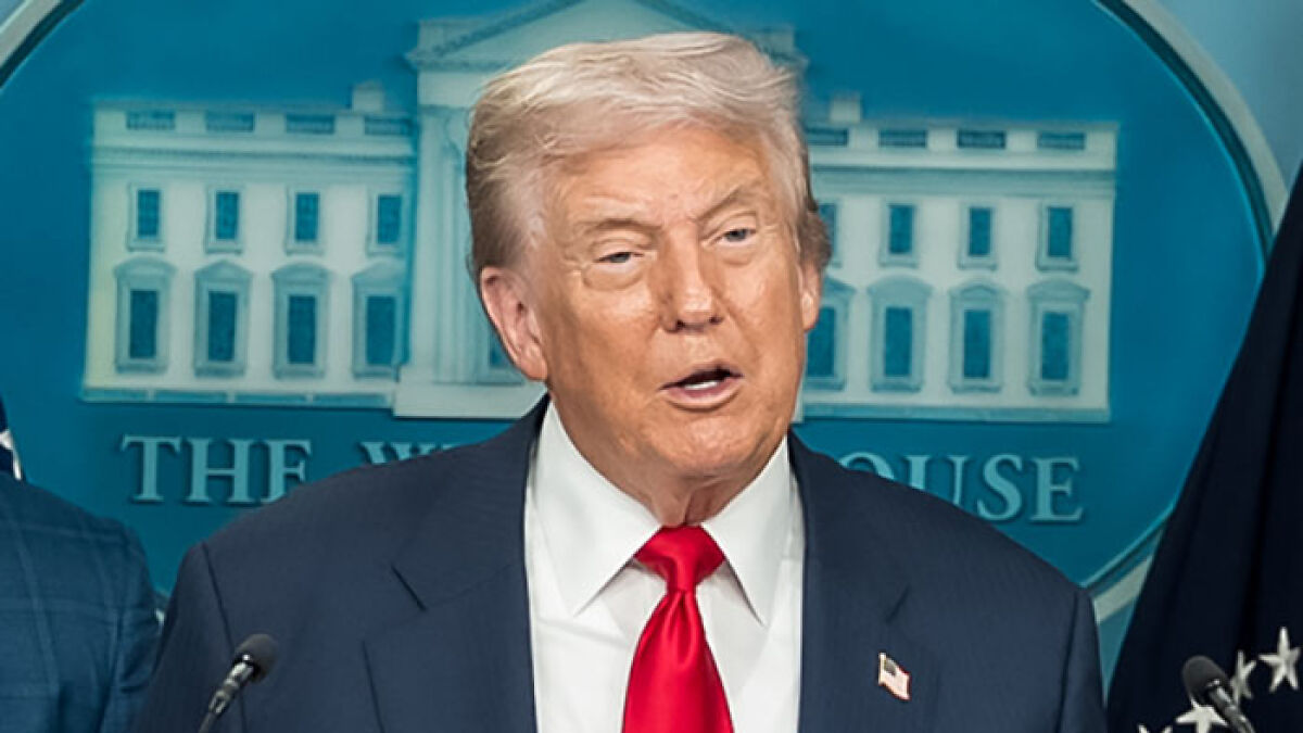 Donald Trump speaking at a press briefing at the White House, wearing a dark suit and red tie.