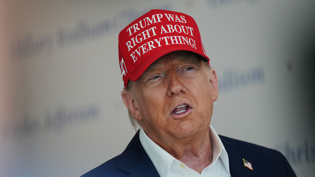 Donald Trump, 79, wearing a red hat, speaking during an event with a serious expression in a formal setting.