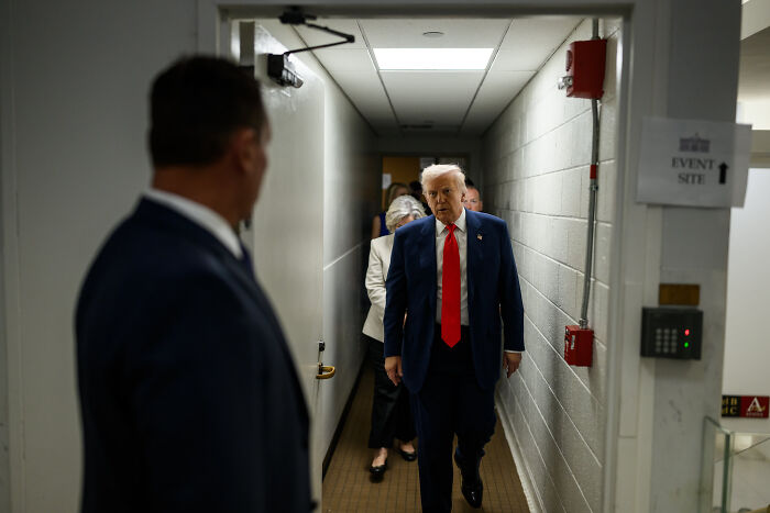 Man in a suit walking down a narrow hallway with security personnel, highlighting secret digital network outing ICE agents.