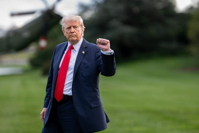 Donald Trump raising fist outdoors, dressed in navy suit and red tie, with background of blurred greenery and helicopter.