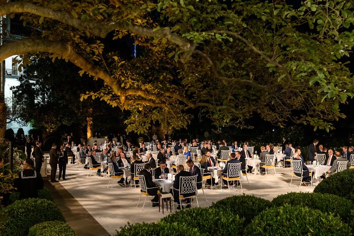 Outdoor formal dinner with guests seated at tables under trees, illustrating Newsom slams Trump’s Rose Garden Club event.
