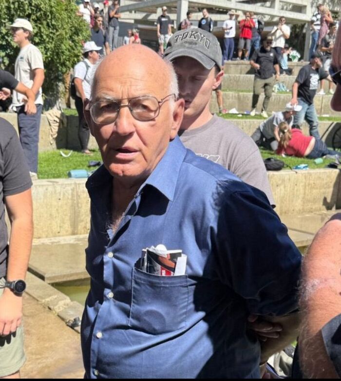 Older man being detained in a crowded outdoor area related to gadfly with disturbing files on his phone, police involved.