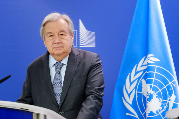 UN official speaking at podium with United Nations flag in background during press event on escalator and teleprompter issues