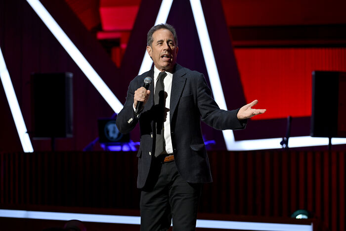 Comedian Jerry Seinfeld performing on stage with microphone, discussing Free Palestine movement and hate group comparison. Comedian Jerry Seinfeld performing on stage with microphone, discussing Free Palestine movement and hate group comparison.