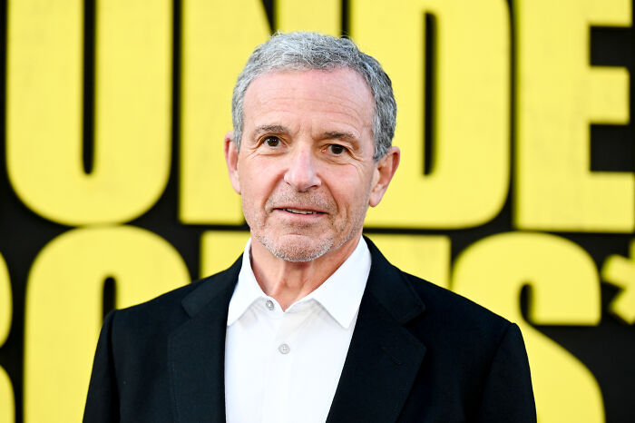 Man in a black blazer and white shirt standing in front of a bright yellow and black backdrop at an event.