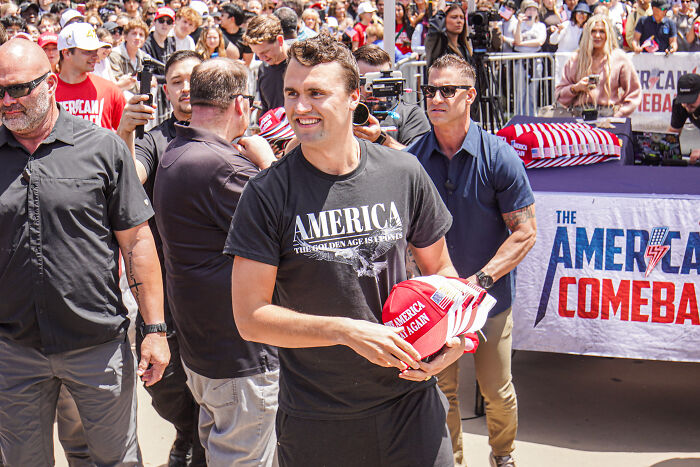 Man holding red hats at a crowded rally highlighting concerns about America’s escalating political violence.