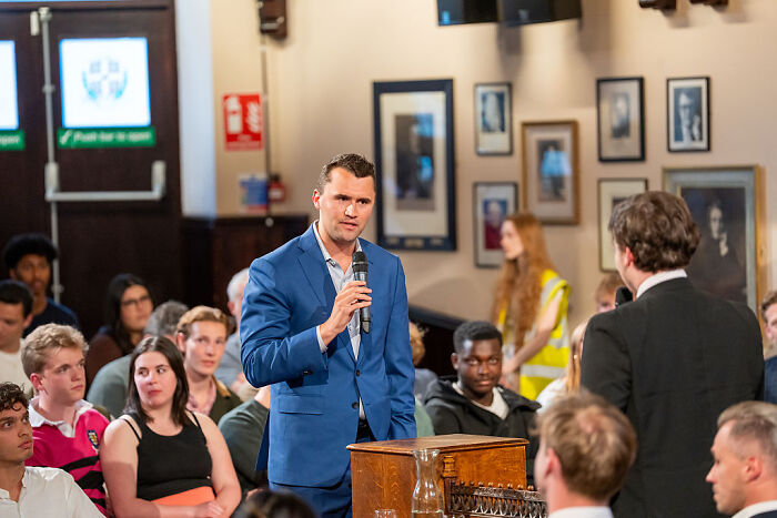 Man in blue suit speaking into microphone at public debate with attentive audience South Park fans call for cancellation