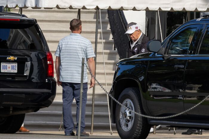 Donald Trump wearing a cap and jacket near black SUVs amid rumors, seen outside with a man and steps behind them.