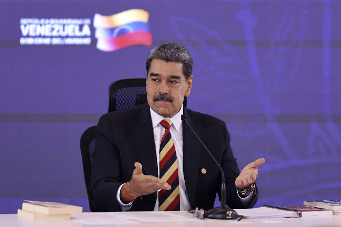 Venezuelan leader speaking at a podium with Venezuela flag backdrop, discussing Trump starting a war with Venezuela.