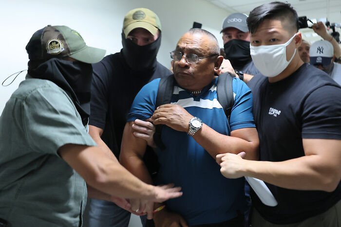 Masked individuals detaining a man in a hallway, illustrating the secret digital network outing ICE agents.