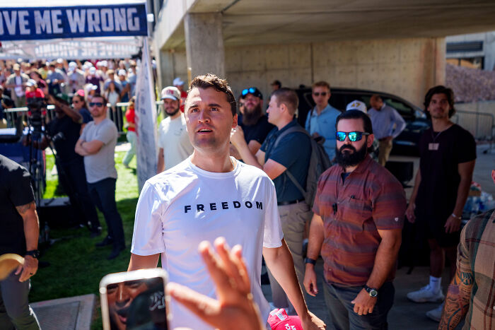 Man wearing a white freedom shirt at a crowded outdoor event with people and security in the background Man wearing a white freedom shirt at a crowded outdoor event with people and security in the background