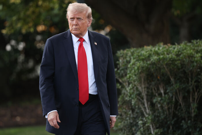 Donald Trump in a dark suit and red tie walking outdoors near green bushes on a cloudy day, discussing illegal substances impact.