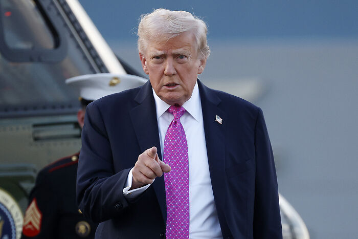 Man in dark suit with pink tie pointing forward outside near a helicopter, discussing left-wing terror movement plans.