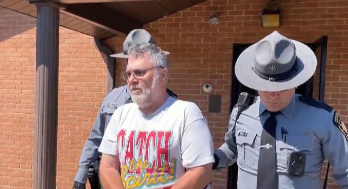 Man in a white graphic t-shirt being escorted by police officers outside a brick building, related to children found in dungeon case