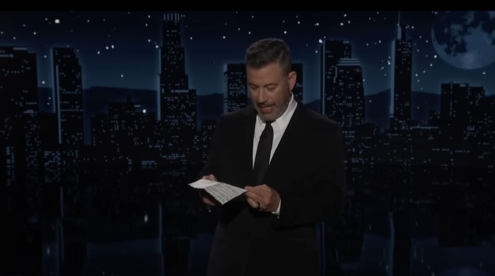 Jimmy Kimmel on a late-night set wearing a black suit, reading notes while mocking Trump in his monologue. Jimmy Kimmel on a late-night set wearing a black suit, reading notes while mocking Trump in his monologue.