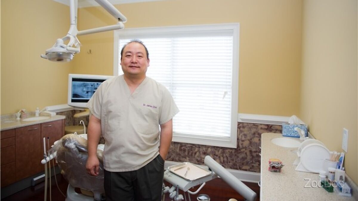 Dentist standing in a dental clinic room with equipment and X-ray on monitor in a well-lit office setting.