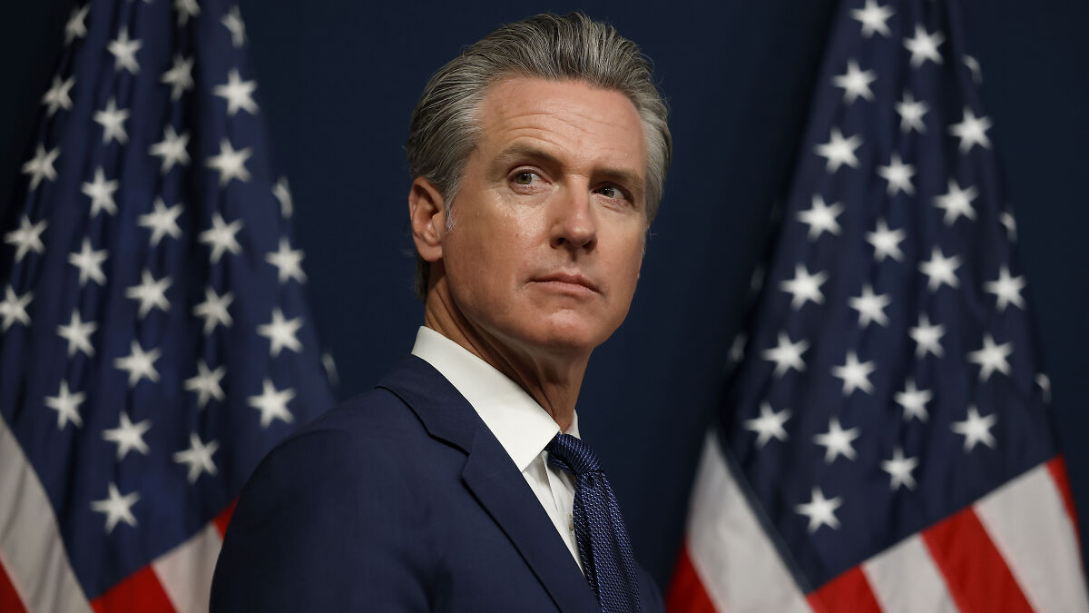 Gavin Newsom in a suit with American flags behind him, addressing media with a serious expression on his face