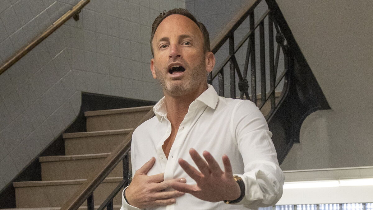 Man in white shirt gesturing with hand raised, appearing surprised, standing near staircase with black railing.