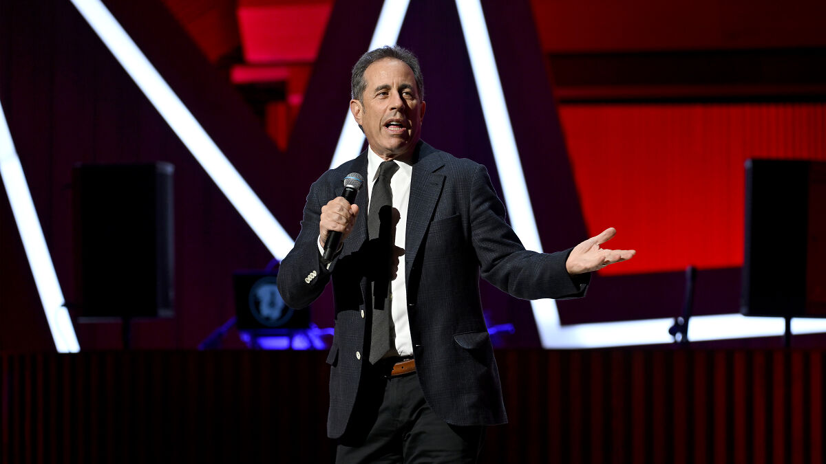 Comedian Seinfeld performing on stage, holding a microphone, discussing Free Palestine movement and hate group comparisons.