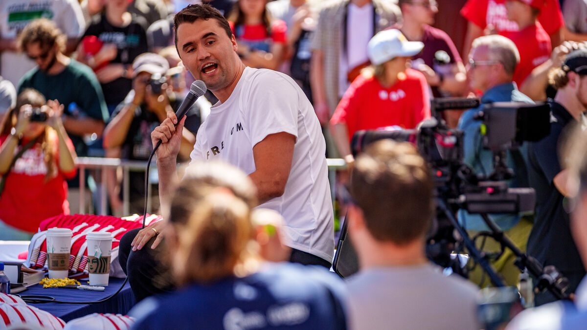 Charlie Kirk speaking into a microphone at an outdoor event with a crowd and media capturing the scene.
