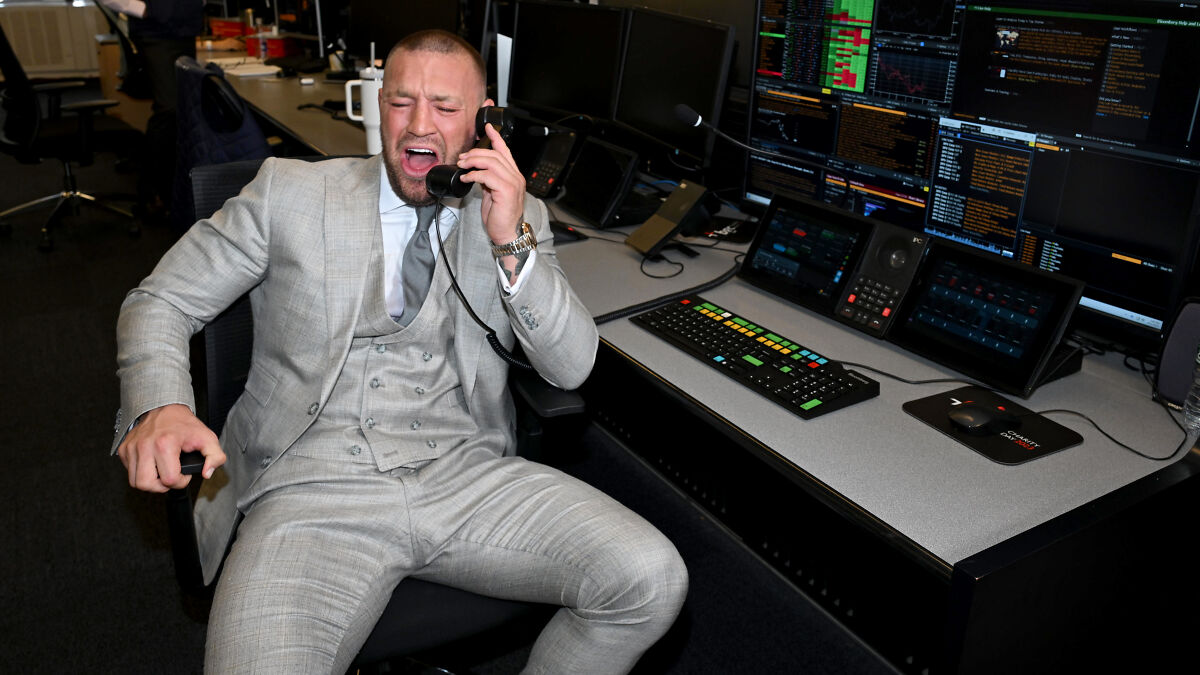 Conor McGregor in a gray suit talking on the phone in a trading office filled with multiple computer screens.