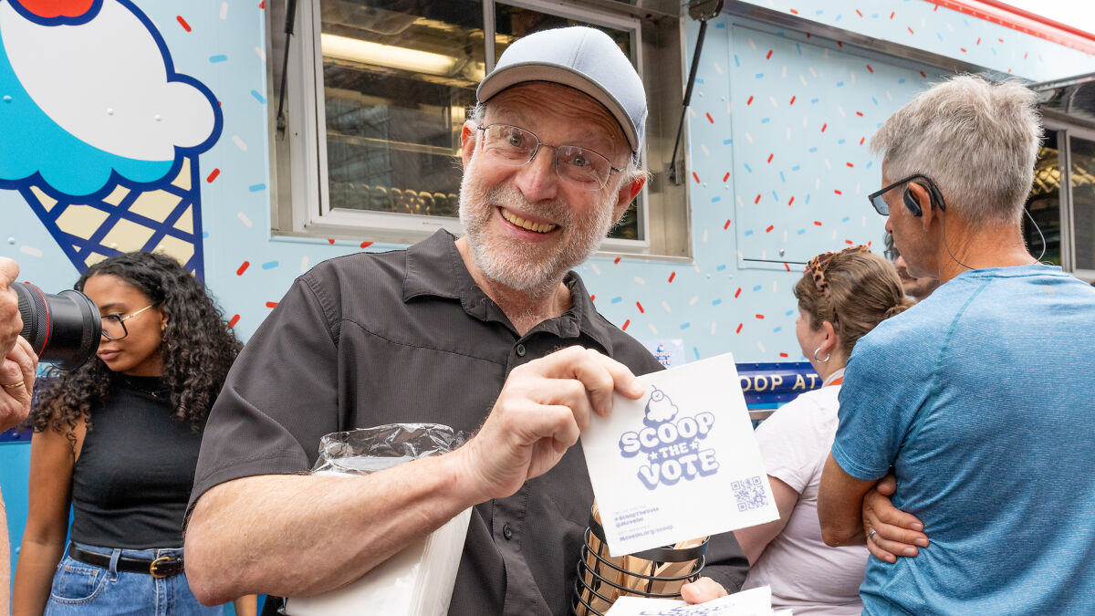 Ben & Jerryu2019s co-founder smiling, holding a Scoop the Vote flyer outside an ice cream truck with people nearby.