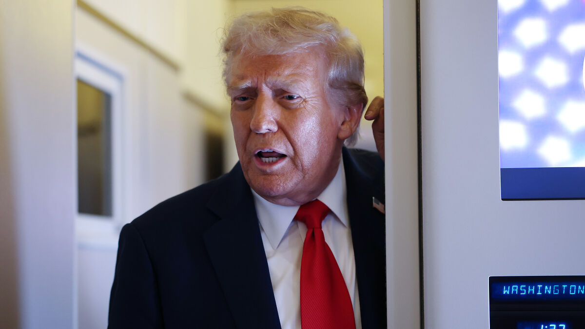 Donald Trump in a suit and red tie speaking near a doorway with glowing screen in Washington location.