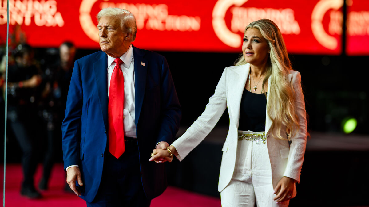 Donald Trump and a woman holding hands at an event, with a red background and focused expressions.