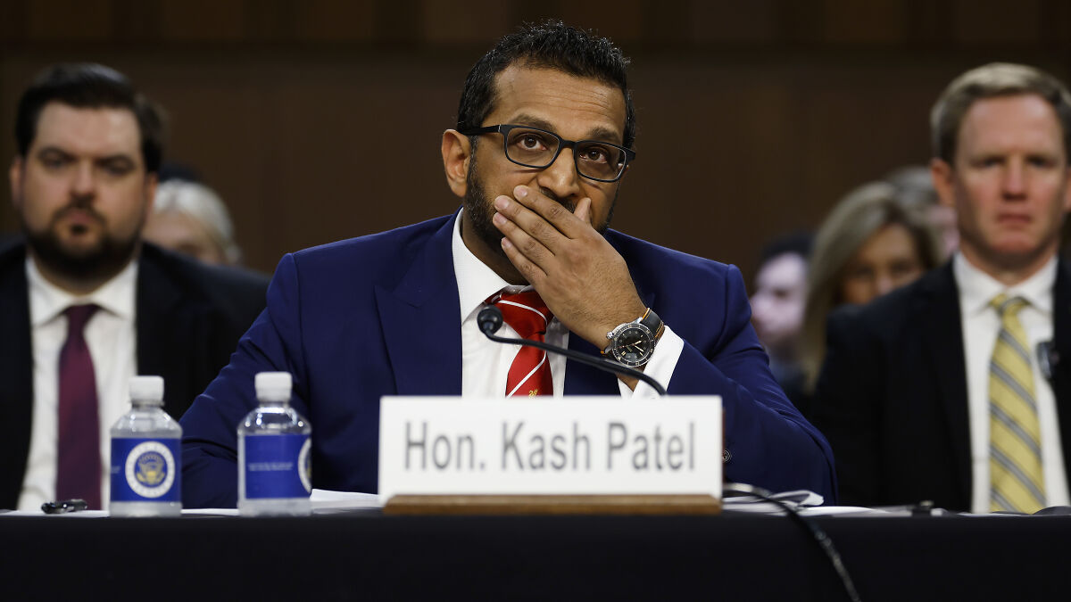 FBI Director Kash Patel in a navy suit and red tie at a hearing, looking thoughtful during a formal discussion.