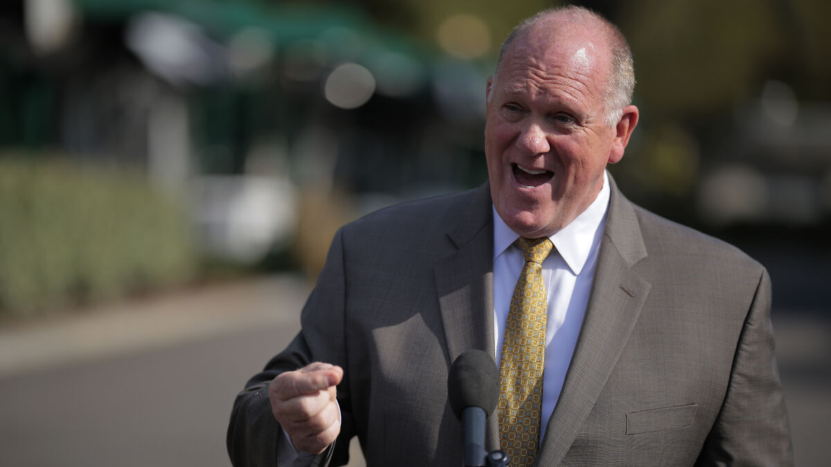 Tom Homan speaking outdoors in a suit and patterned yellow tie, discussing border issues near a microphone.