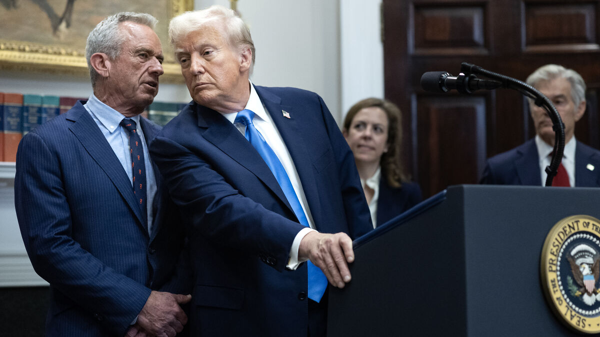 Donald Trump in a navy suit speaking privately with a man in a pinstripe suit, discussing autism and Tylenol claims at a podium.