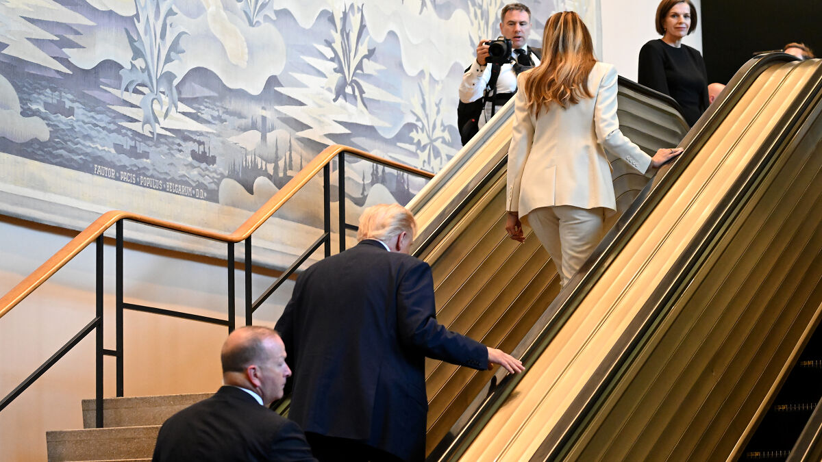 Trump and Melania ascending escalator inside UN building as photographers and officials observe nearby