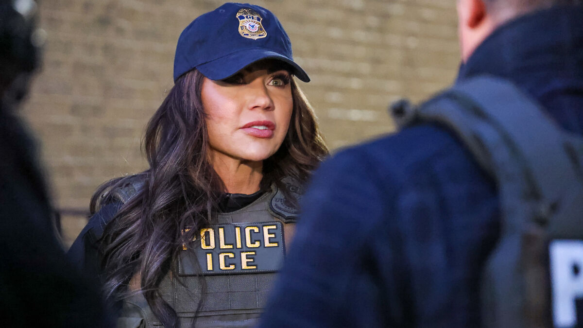 Woman wearing an ICE police vest and cap, talking to colleagues during a law enforcement activity in an urban setting.