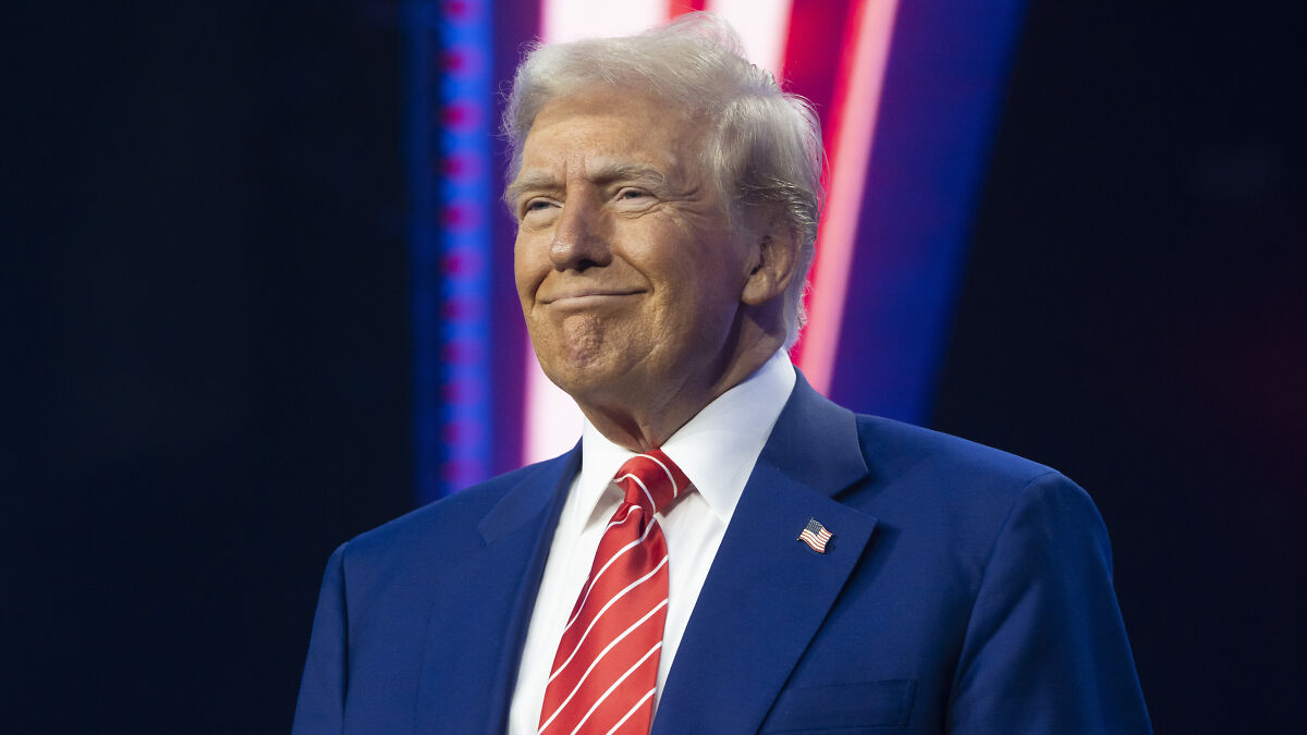 Former President Trump wearing a blue suit and red striped tie at an event with colorful lighting in the background