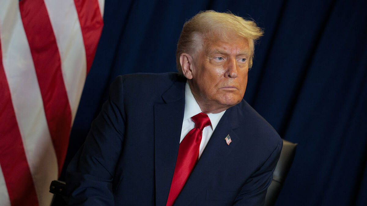 Donald Trump in a navy suit and red tie, looking to the side with an American flag in the background during a speech event.