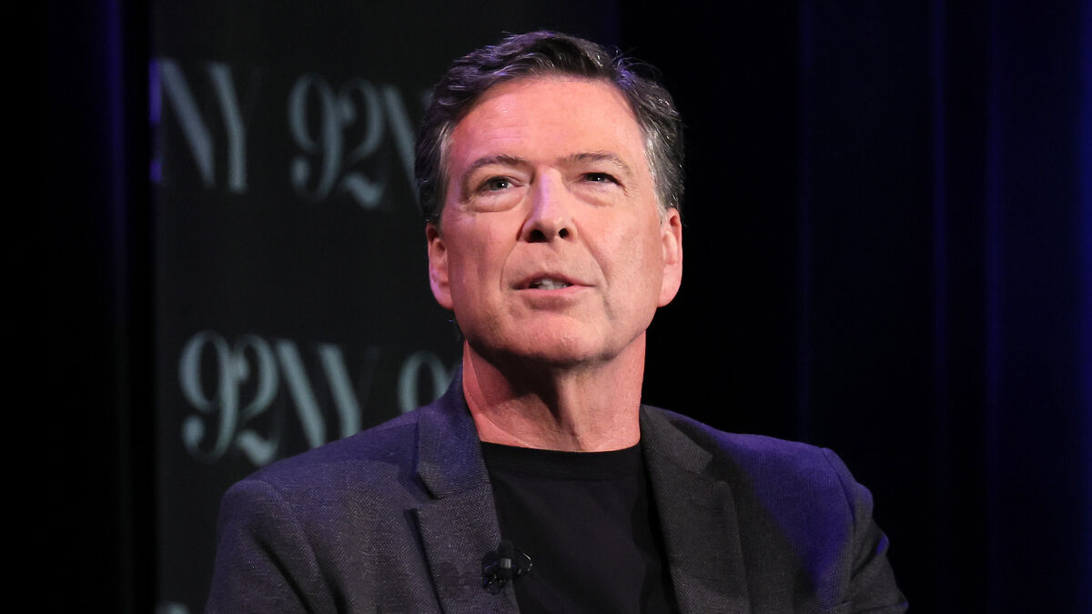 Former FBI chief James Comey speaking at an event, wearing a dark jacket and black shirt against a dark background.