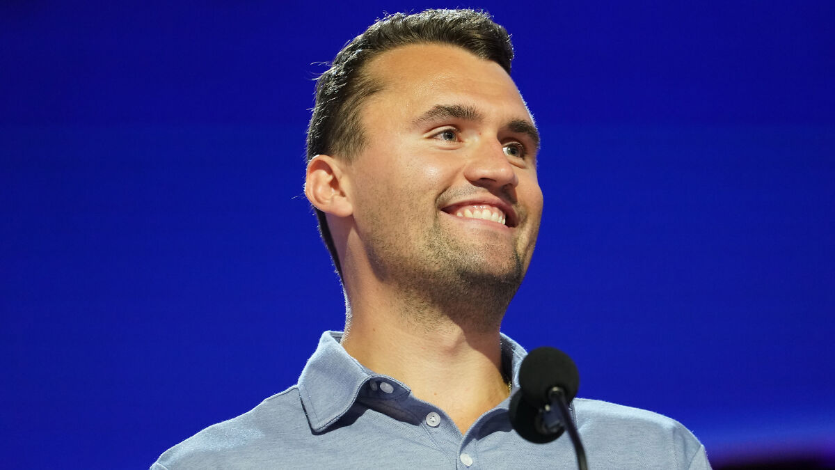 Charlie Kirk smiling at a podium with a microphone against a blue background in a public speaking event