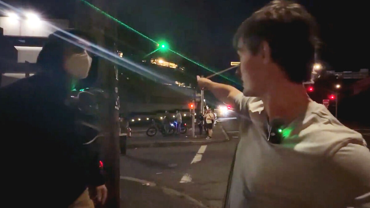 Man points at green laser beam at night during Portland protest, with DHS snipers allegedly aiming lasers at Antifa member.