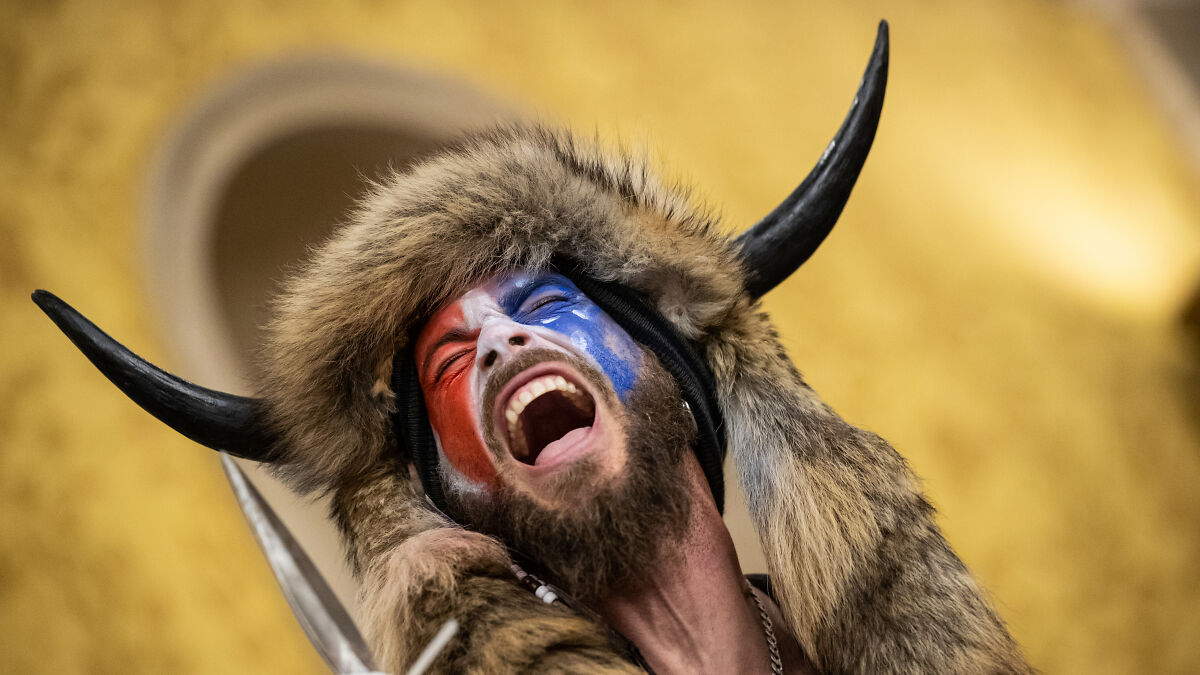 QAnon Shaman wearing fur hat with horns and painted face shouting in a dramatic and intense moment.