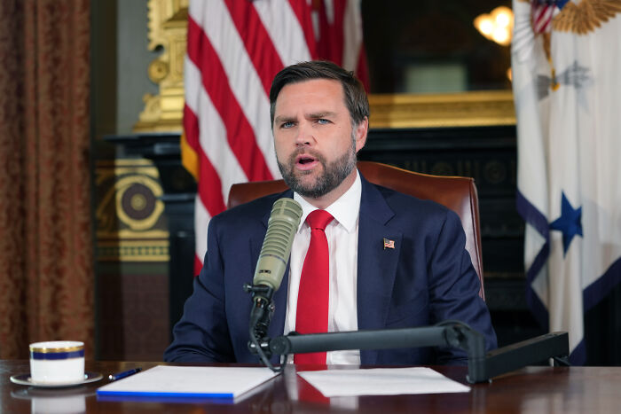 JD Vance speaking at a desk with microphone, urging public to report people celebrating Charlie Kirk shooting incidents.