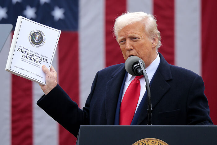 Donald Trump speaking at a podium holding a report on foreign trade barriers against the backdrop of an American flag discussing trade war costs.