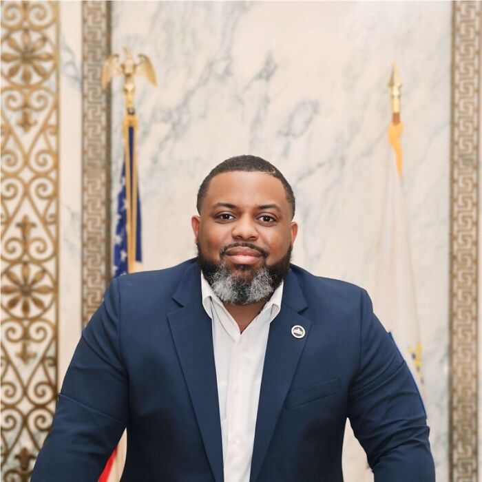 Man in navy suit with white shirt posing in an official government office setting with flags in the background. Man in navy suit with white shirt posing in an official government office setting with flags in the background.