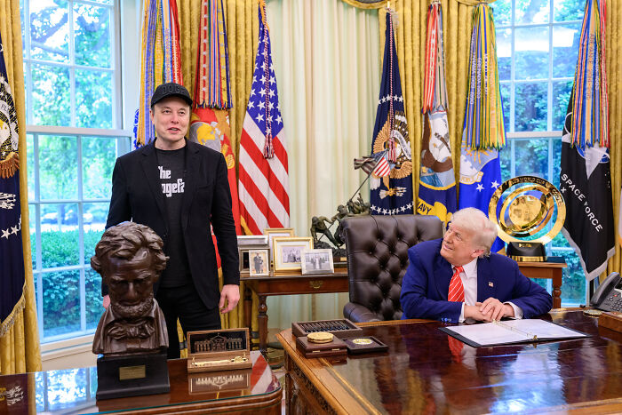 Elon Musk standing beside a bust in an office while a man in a suit looks on, featuring flags and official decor.